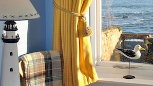 Una habitación con vista al mar, una lámpara, un sillón con manta escocesa y una figurita de gaviota sobre un pedestal.