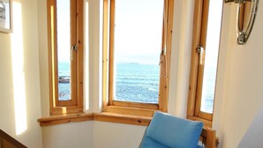 Una habitación con ventana de madera, una silla azul con cojín escocés y una mesa de madera con un libro.