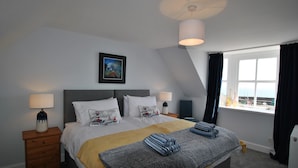 Cama bem arrumada com manta listrada em cinza e azul, mesa de cabeceira de madeira com abajur e quadro na parede.