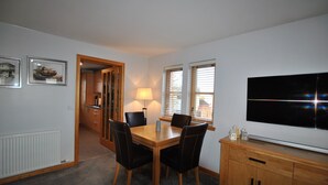 Een eetruimte met een houten tafel en stoelen, een lamp, een televisie op een houten kast, en een deur die naar een andere kamer leidt.