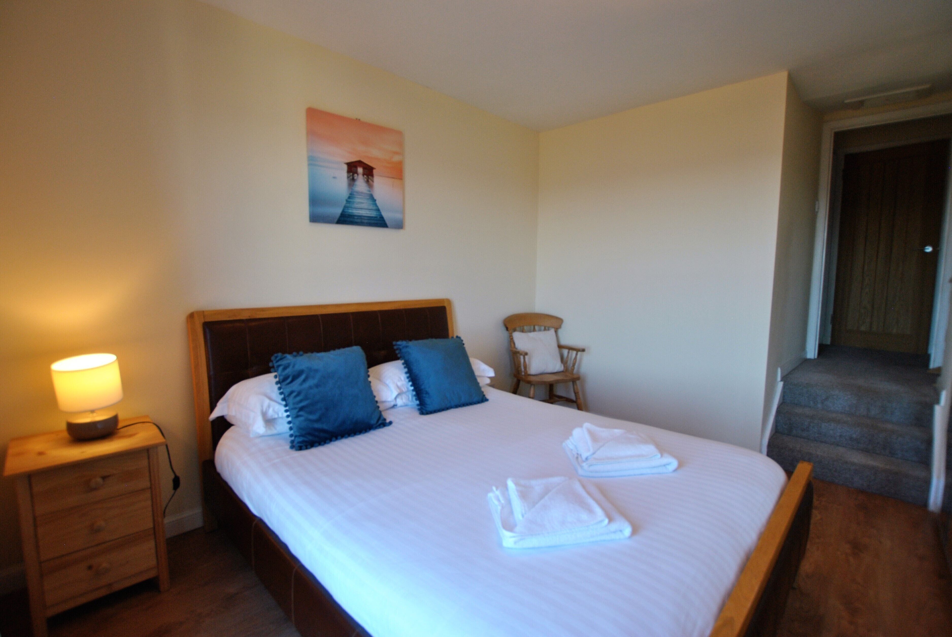 Een net opgemaakt bed met kussens, een nachtkastje met een lamp, een stoel en een schilderij aan de muur.