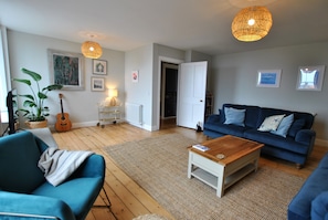 A living room with a blue sofa, a wooden coffee table, a blue armchair, a guitar, a plant, and a wicker lamp.