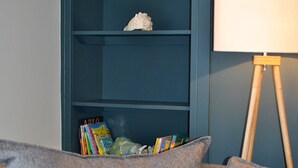 Un sofá gris con cojines estampados, una estantería con libros y objetos decorativos, y una lámpara de pie.