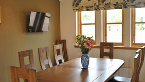 Ein Essbereich mit Holztisch, Stühlen, einer Vase mit Blumen, einem Fernseher an der Wand und Fenstern mit Vorhängen.