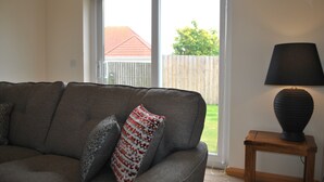 Ein Wohnzimmer mit grauem Sofa, einem gemusterten Kissen, einer Lampe auf einem Holztisch und Blick durch eine Glastür in einen Garten.