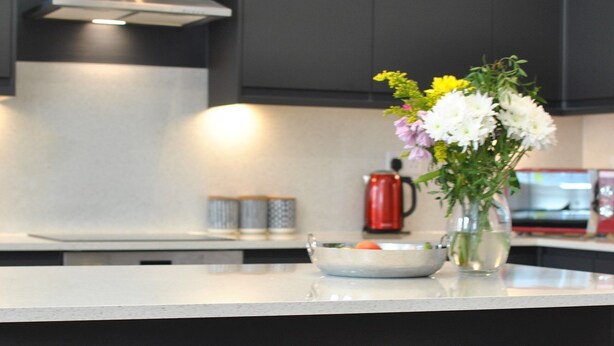 Een moderne keuken met donkere kastjes, een roestvrijstalen afzuigkap, een wit aanrechtblad en twee witte barkrukken met houten poten.