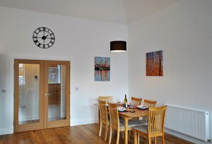 A dining area with a wooden table set for four, surrounded by chairs, a clock on the wall, and two paintings. - Academy Apartment Anstruther- stunning luxury home (Anstruther)