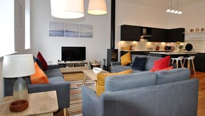 Een moderne woonkamer met een grijze zithoek, een flatscreen televisie en een houten salontafel. Achtergrond is een keuken zichtbaar met donkere kastjes en een centraal kookeiland.