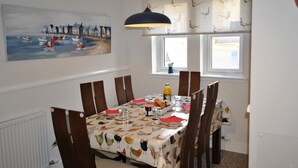 A dining area with a table set for four, a painting of boats on the wall, and a window with curtains.