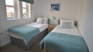 A bedroom with two single beds, a bedside table with a lamp, a window with a view of the outside, and a painting on the wall.