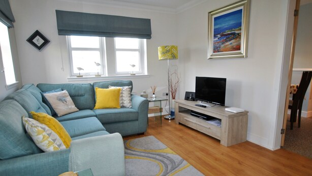 A living room with a teal sofa, a wooden coffee table, a flat-screen TV on a wooden stand, and a painting on the wall.