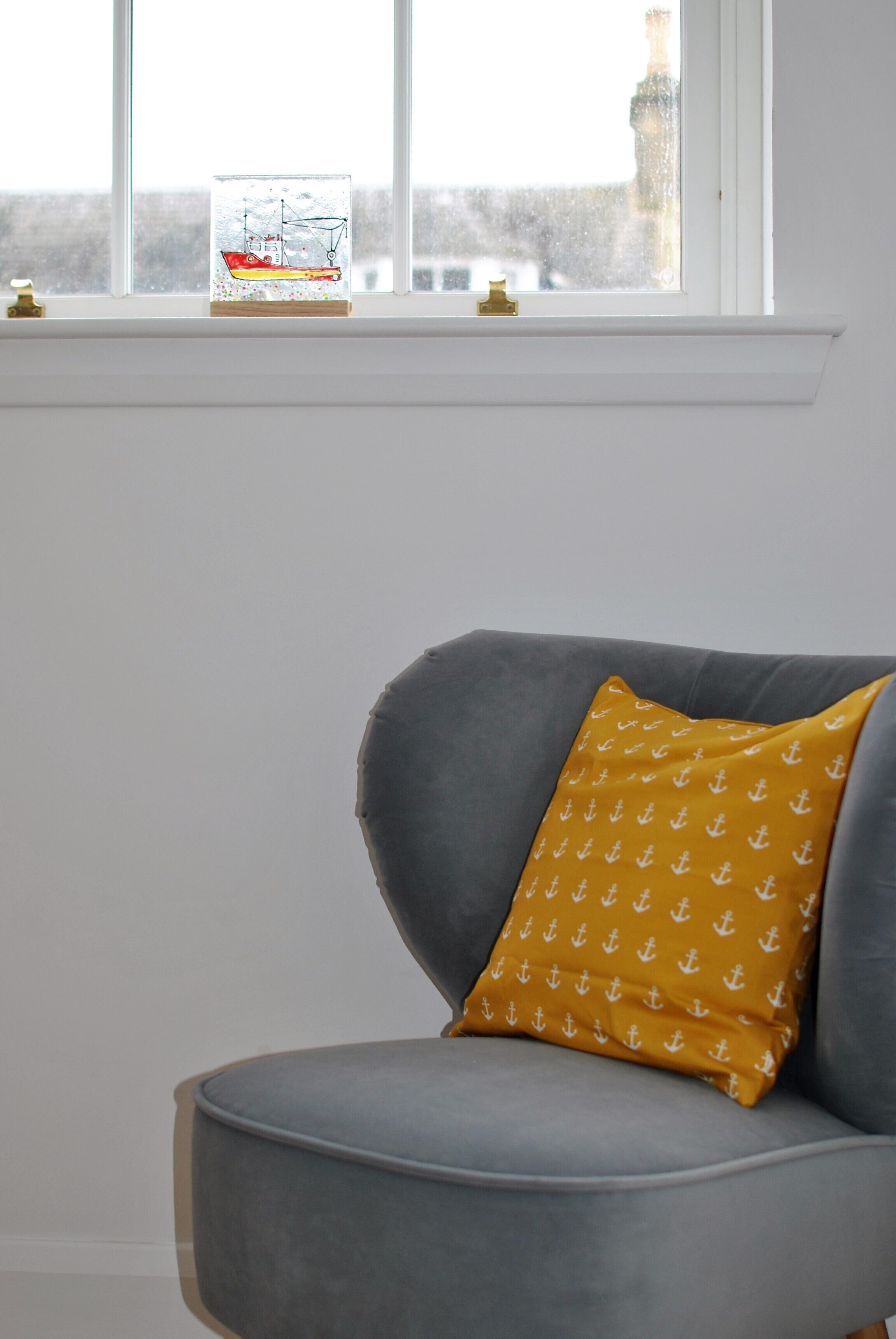 Un fauteuil gris agrémenté d’un coussin jaune moutarde orné de motifs d’ancres, placé devant une fenêtre offrant une vue dégagée sur un bâtiment à l’extérieur.
