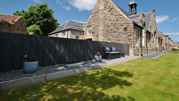 A stone building with a steep roof and a bell tower, surrounded by a well-maintained lawn and a wooden fence.