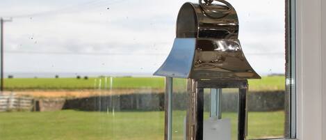 Un farol de metal sobre una superficie blanca, cerca de una ventana con vistas a un campo verde.