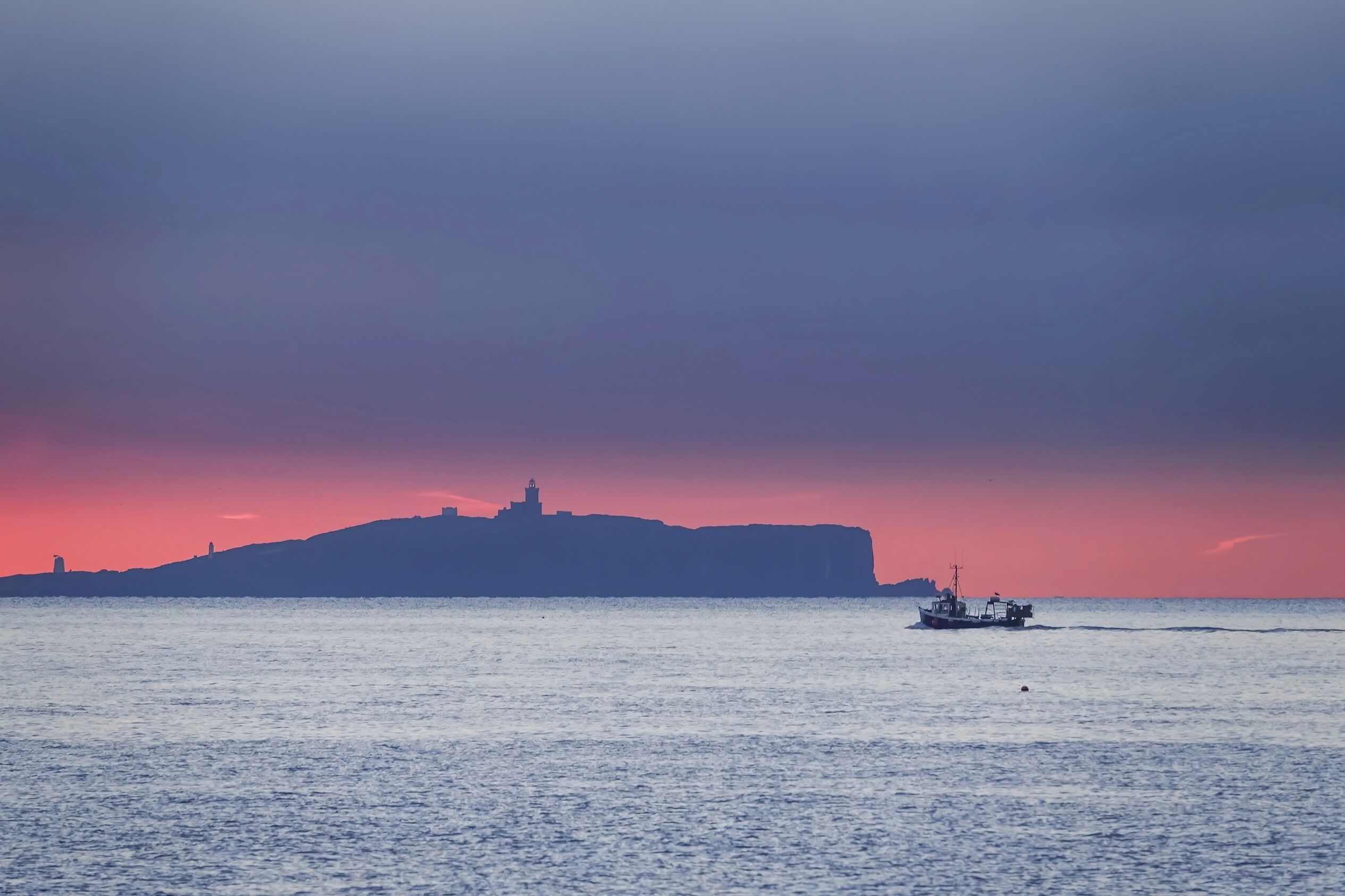 A coastal scene with a boat on calm waters, a distant hill with a structure, and a sunset sky.