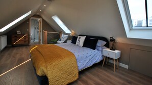 Et soveværelse med skrå vægge, en seng med et mønstret sengetæppe, et sengebord med en lampe, og et vindue der lukker naturligt lys ind.