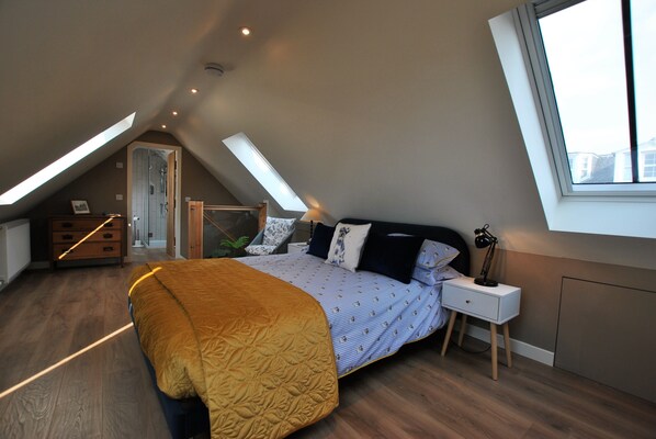 A bedroom with a sloped ceiling, a bed with a patterned bedspread, a bedside table with a lamp, and a window providing natural light.