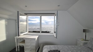 Et soveværelse med et stort vindue der vender ud mod havet, en seng med et mønstret sengetæppe, et natbord med en lampe og en hvid kommode.