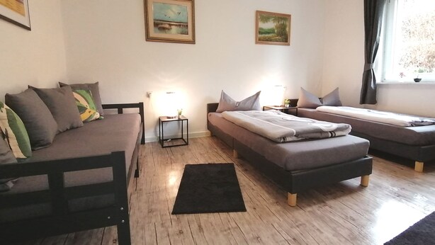 Una habitación con un sofá, dos camas individuales, una mesita con una lámpara y dos cuadros en la pared.