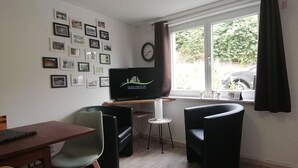 Una habitación con televisión sobre un mueble bajo, una mesa de comedor, dos sillas, un reloj y una ventana con cortinas.
