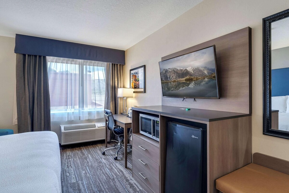 A hotel room with a bed, desk, air conditioning unit, television, and a painting on the wall.