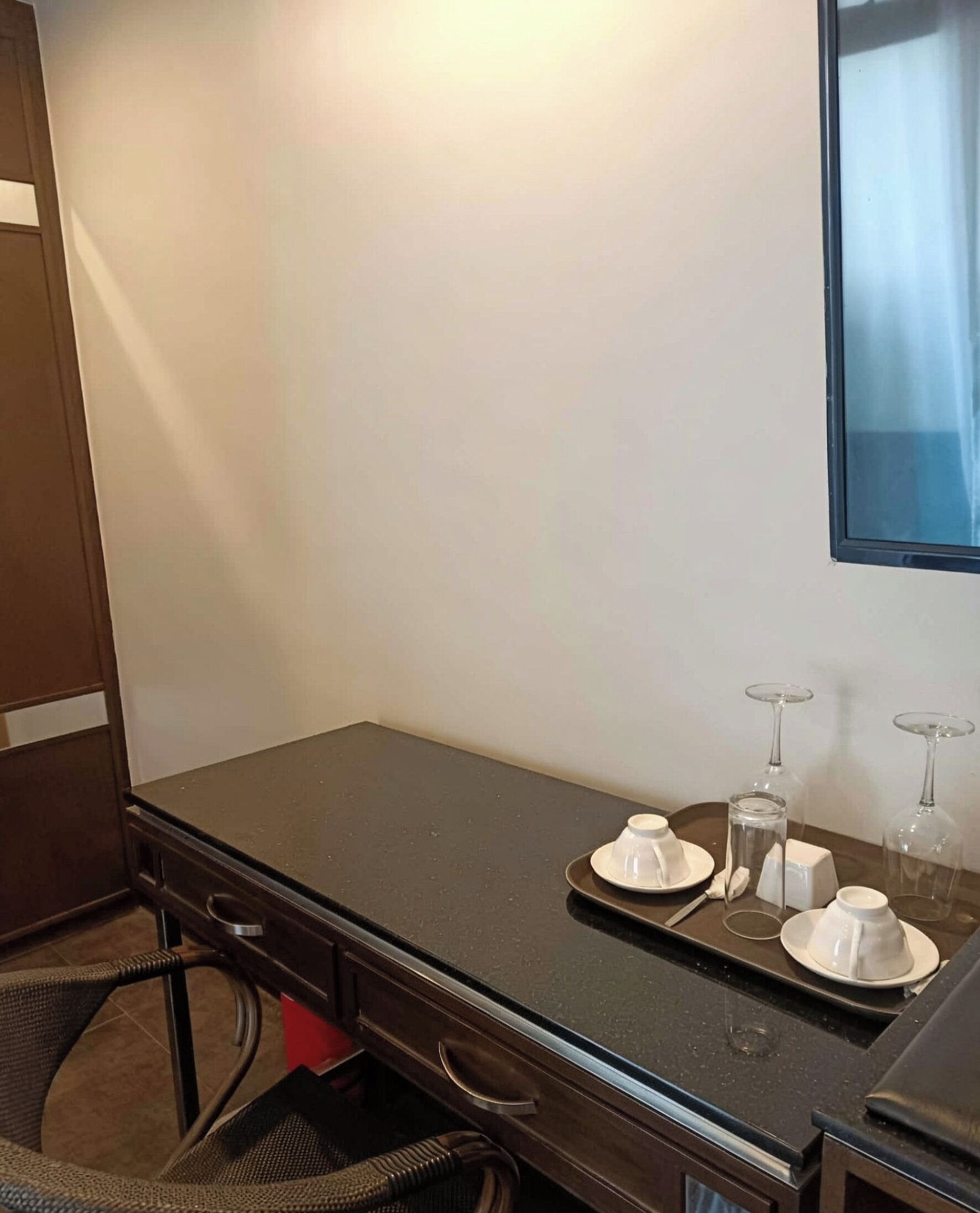 A hotel room with a dark wooden desk, a chair, a small table with a tray holding a teapot, cups, and wine glasses.