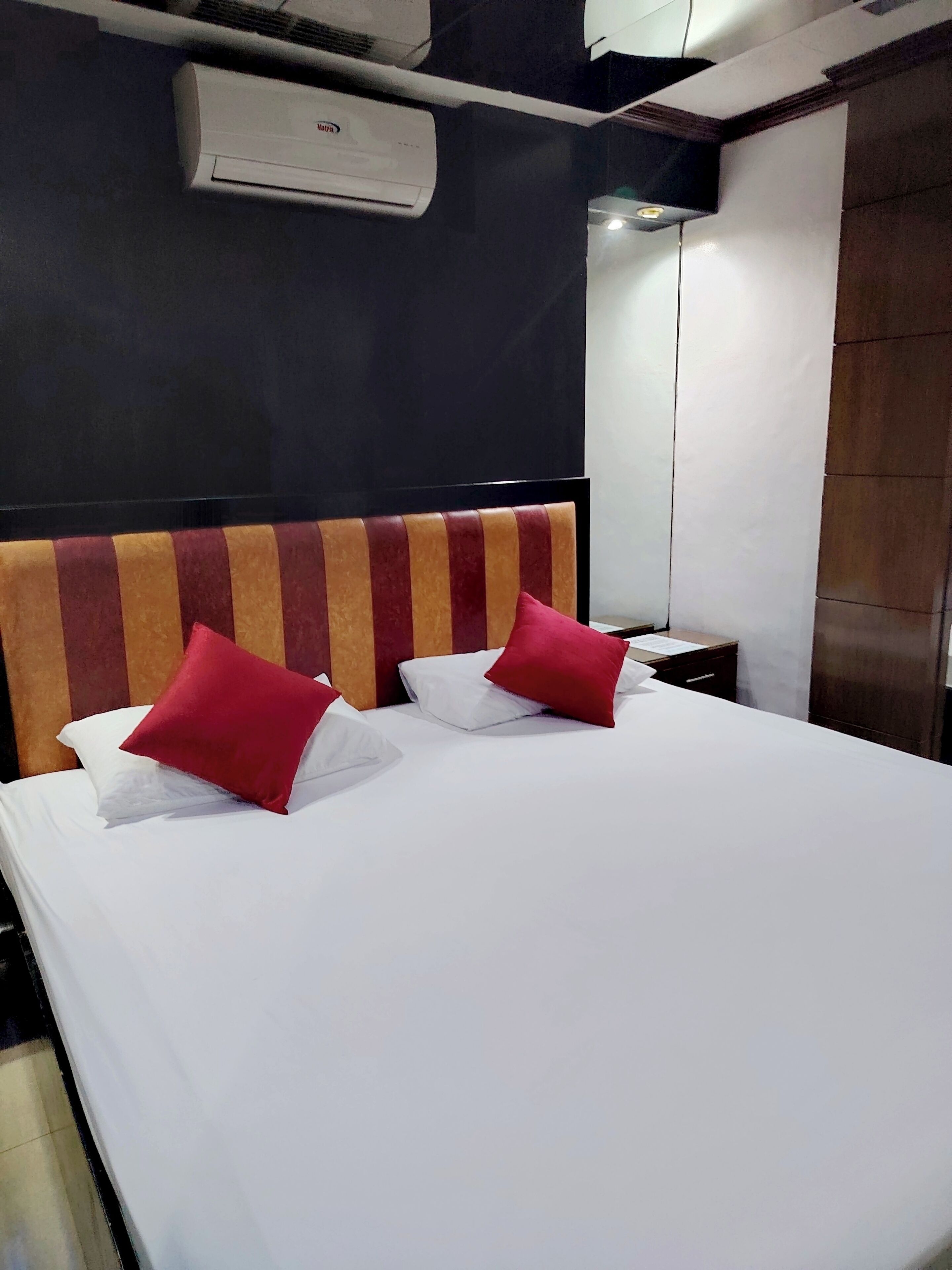 A hotel room with a bed, two pillows, a nightstand, a mirror, and an air conditioning unit.