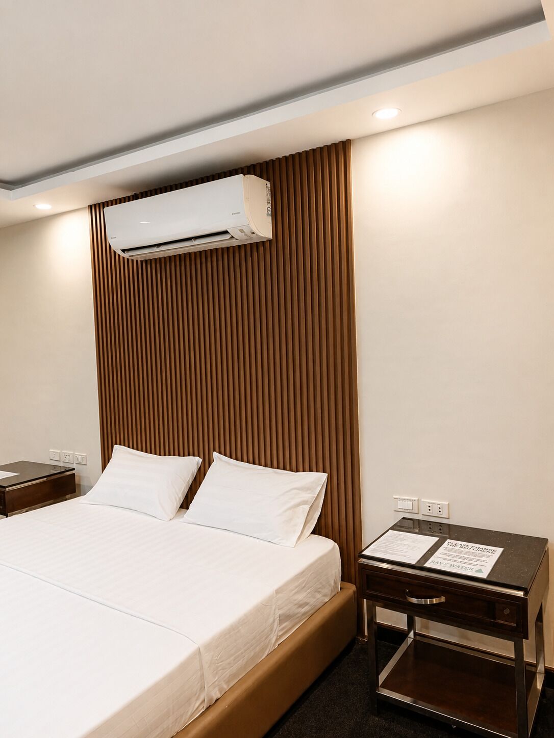 A hotel room with a bed, a nightstand, an air conditioning unit, and a water conservation sign.