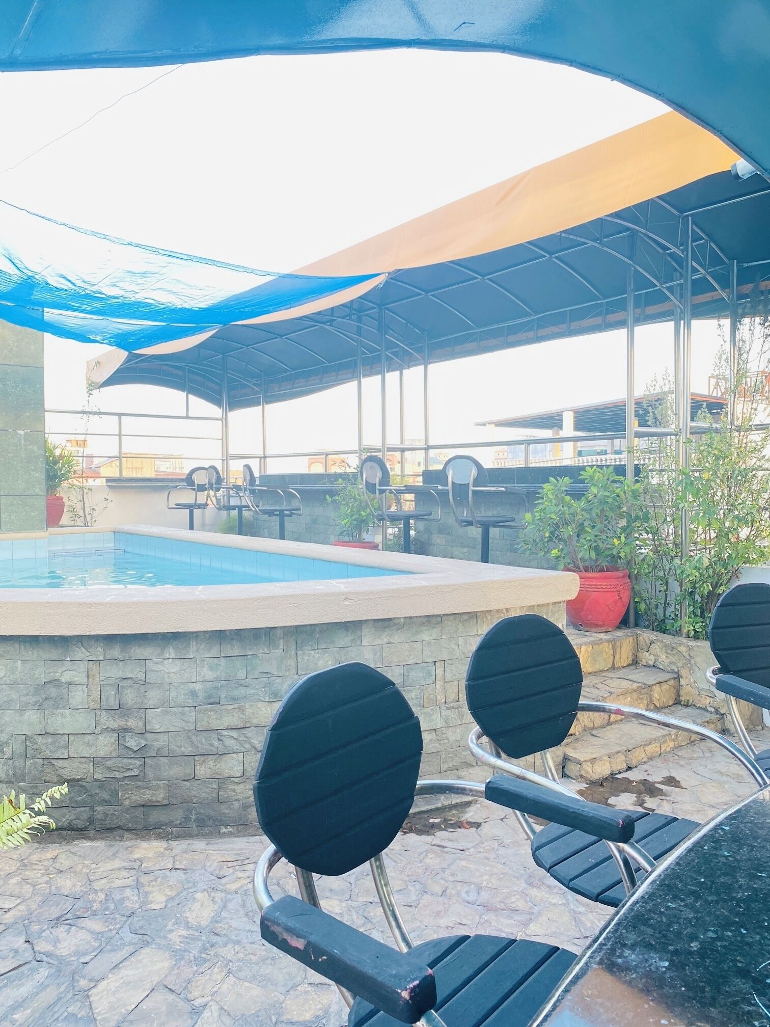 An outdoor pool area with stone walls, black lounge chairs, and a covered seating area with umbrellas.