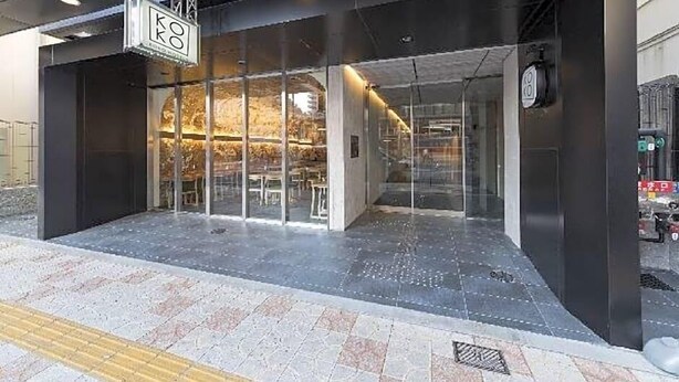 A modern building entrance with glass doors, a visible interior with tables and chairs, and a sign above the entrance.