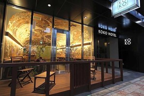 L’extérieur de l’hôtel Koko se caractérise par de grandes fenêtres en verre, une terrasse en bois avec des sièges et un panneau indicateur.
