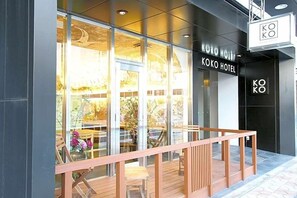 Une entrée d’hôtel moderne avec des portes en verre, une terrasse en bois et une enseigne indiquant « HÔTEL KOKO ».