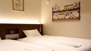 Een net opgemaakt bed met witte lakens en handdoeken, een nachtkastje met een klok, en een wandplank met borden.