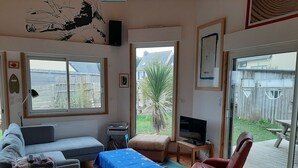 A living room with a sofa, a small table, a TV, and a chair. There are large windows and a view of the outside.