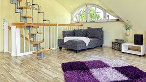 Ein modernes Wohnzimmer mit einer Holztreppe, einem grauen Sofa, einem violetten Teppich und einem weißen Konsolentisch.