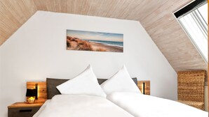 Un dormitorio con techo inclinado, una cama con ropa de cama blanca, cabecera de madera, mesita de noche con lámpara y una pintura de una escena de playa en la pared.