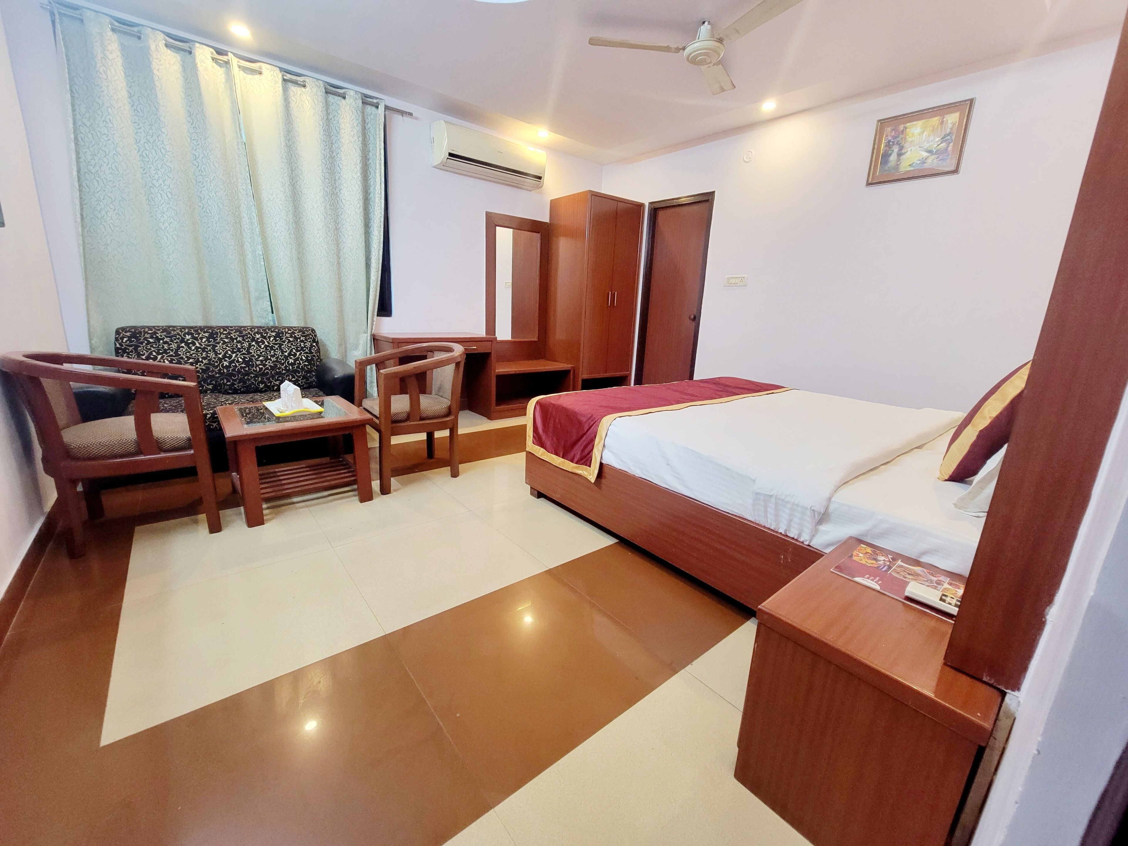 A hotel room with a bed, a desk, two chairs, a sofa, an air conditioning unit, a ceiling fan, and a window with curtains.