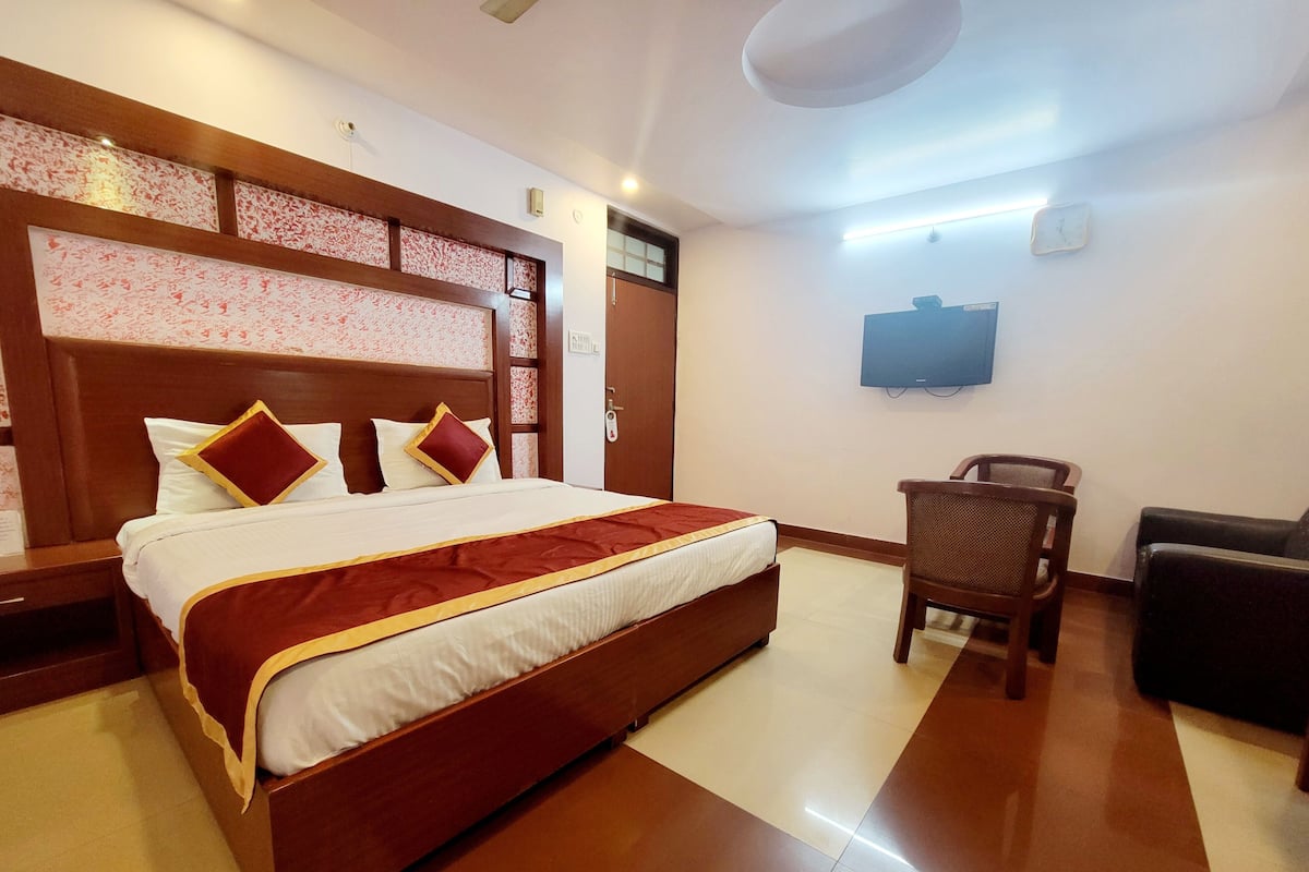 A hotel room with a large bed, a wooden headboard, a flat-screen TV mounted on the wall, a wooden chair, and a sofa.