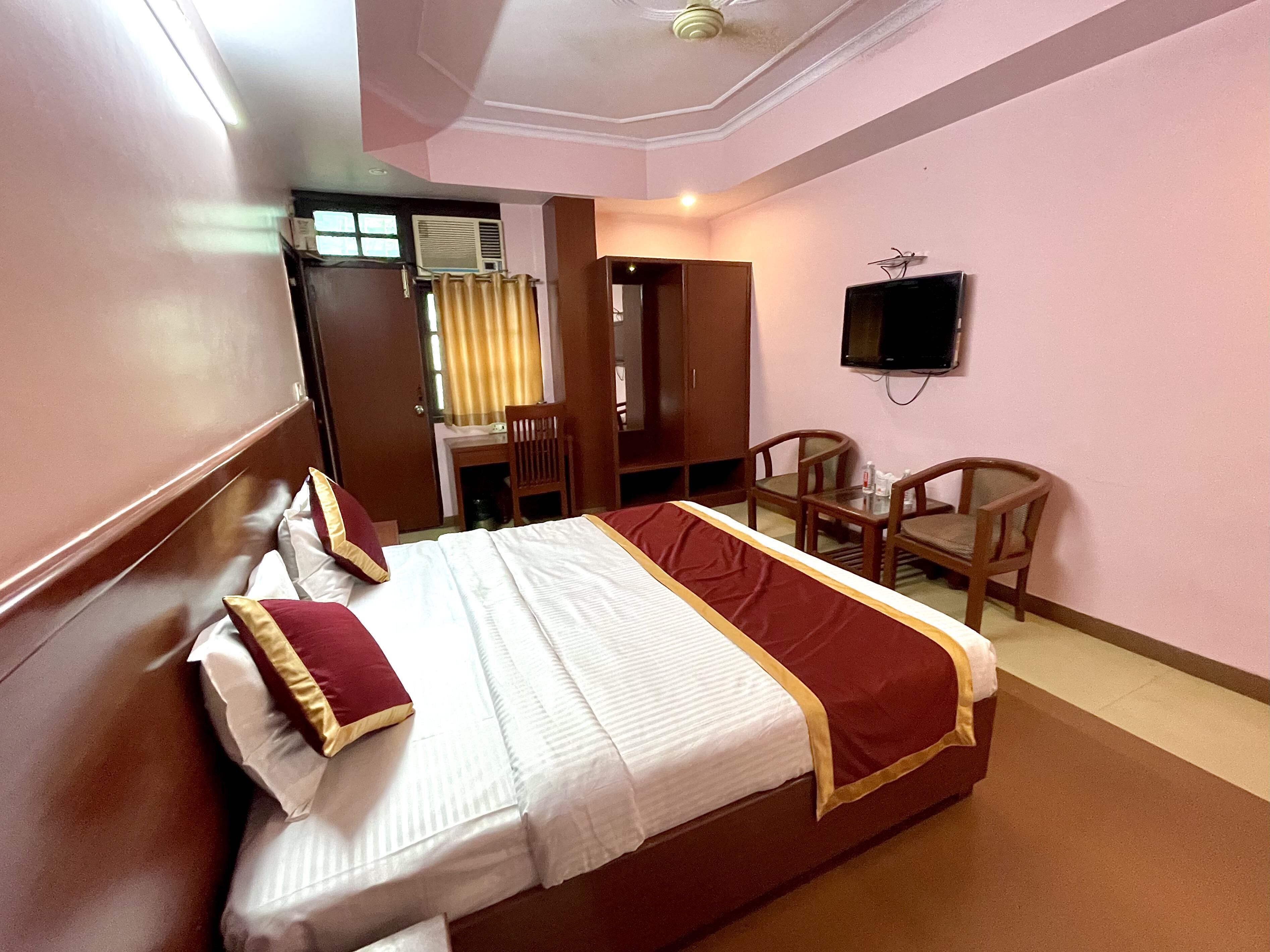 A hotel room with a large bed, a desk, two chairs, a television, and an air conditioning unit.