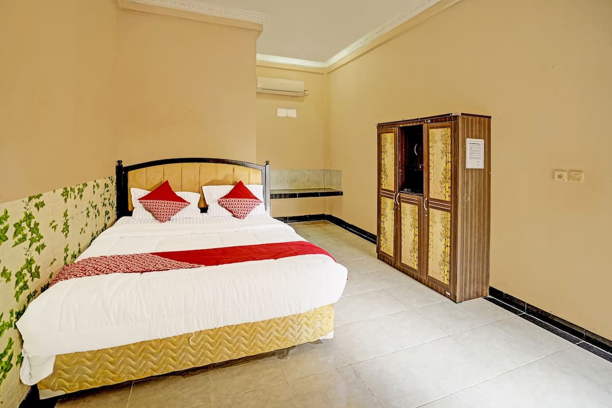 Un lit bien fait, recouvert d’une couverture blanche et rouge, avec une tête de lit en bois et deux oreillers rouges. Un meuble en bois avec des portes en verre se trouve à droite du lit.