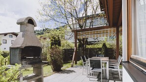 A stone-built outdoor oven, a patio with chairs, and a view of a tree and buildings in the background. - Chalet Badia by we rent (Zell am See)