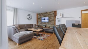 A modern living room with a sectional sofa, a coffee table, a TV mounted on a stone wall, and a wooden dining table. - Chalet Badia by we rent (Zell am See)