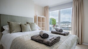 Una cama bien hecha con toallas enrolladas, cabecera y vistas a un paisaje urbano a través de amplios ventanales.