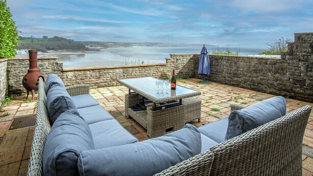 Un patio con mobiliario de mimbre, una mesa con vasos y una botella, y vista al mar.