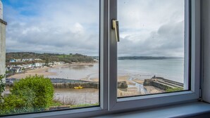 Vista costera a través de una ventana, con playa, muro de contención y una línea de costa distante con edificios.