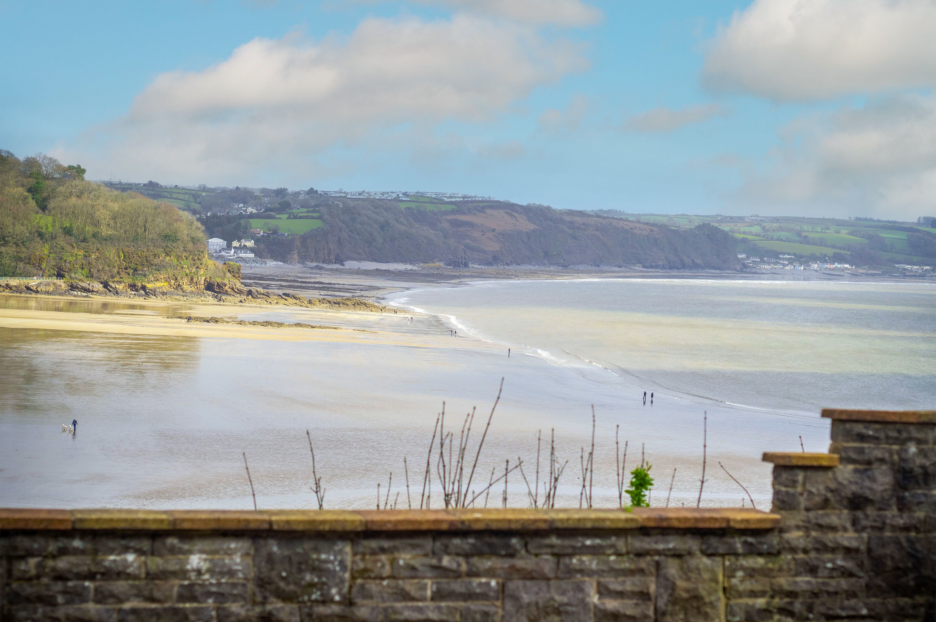 A coastal view with a stone wall in the foreground, a sandy beach, and a hilly landscape in the background.