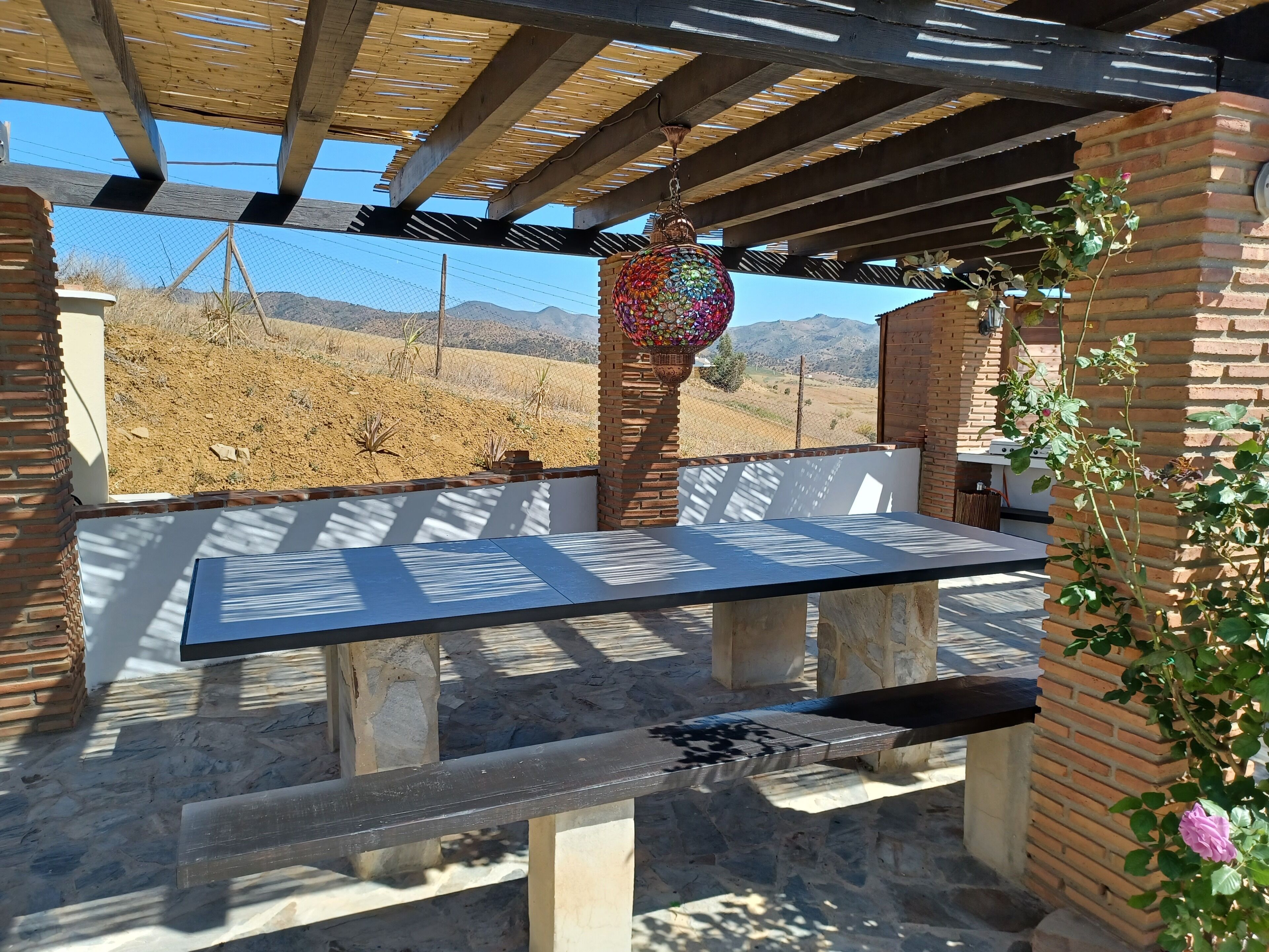 An outdoor patio with a table and benches, a hanging decorative item, and a view of a hilly landscape.