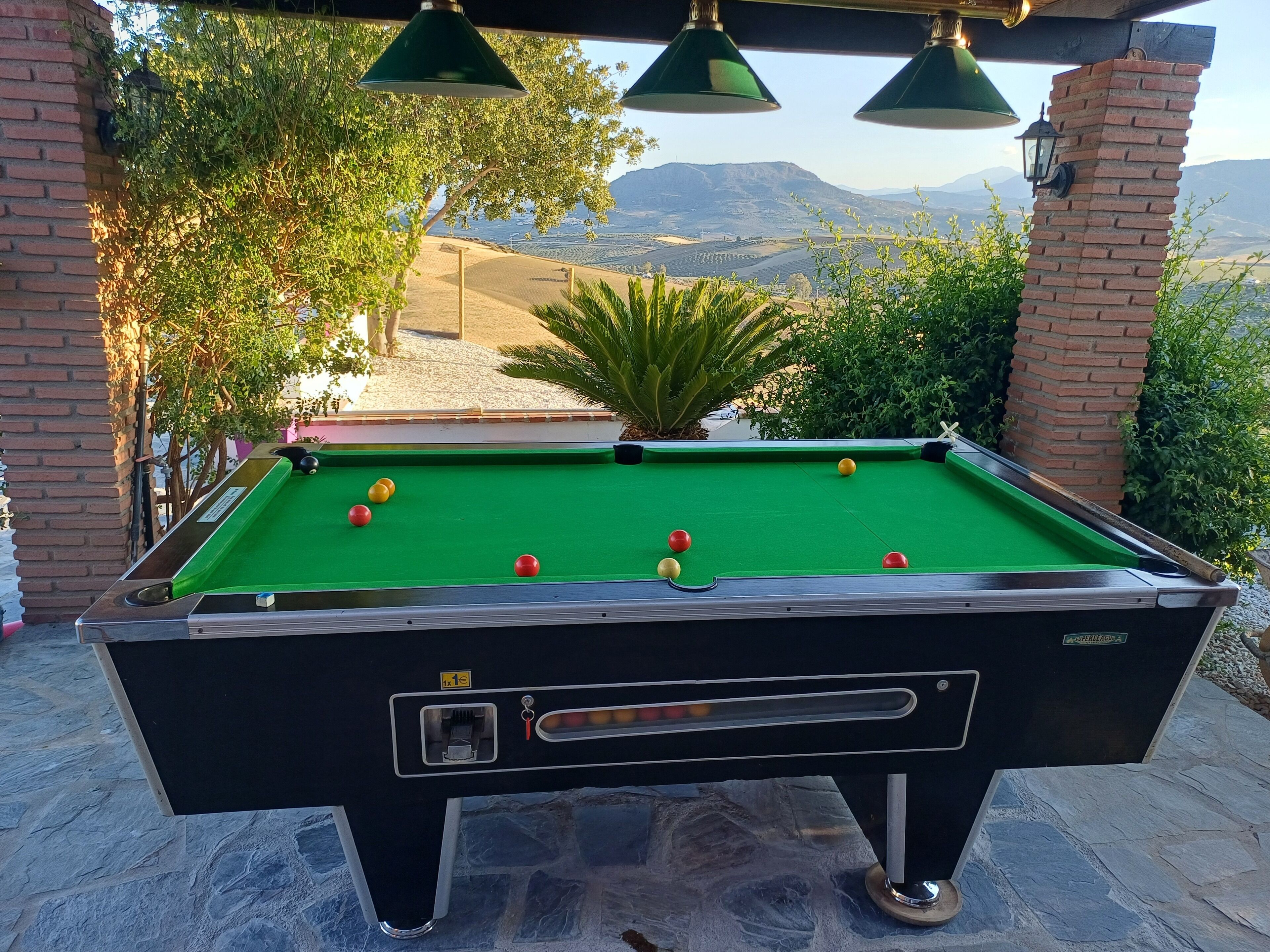 Une table de billard avec ses billes, entourée de piliers en briques et de lampes vertes, le tout mis en valeur par un paysage pittoresque de montagnes et d’arbres.