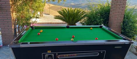 Une table de billard avec ses billes, entourée de piliers en briques et de lampes vertes, le tout mis en valeur par un paysage pittoresque de montagnes et d’arbres.