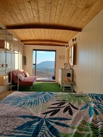 Ein Schlafzimmer mit einem großen Bett, einem roten Sessel, einem Schreibtisch und Blick auf eine gebirgige Landschaft durch eine Schiebetür.
